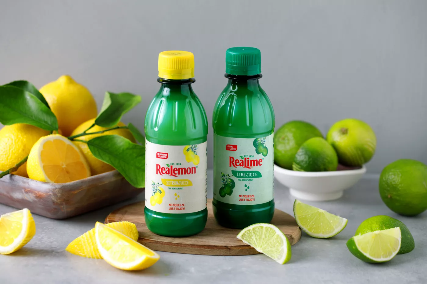 To flasker med sitron- og limedråper står på et bord med sitroner og lime rundt.